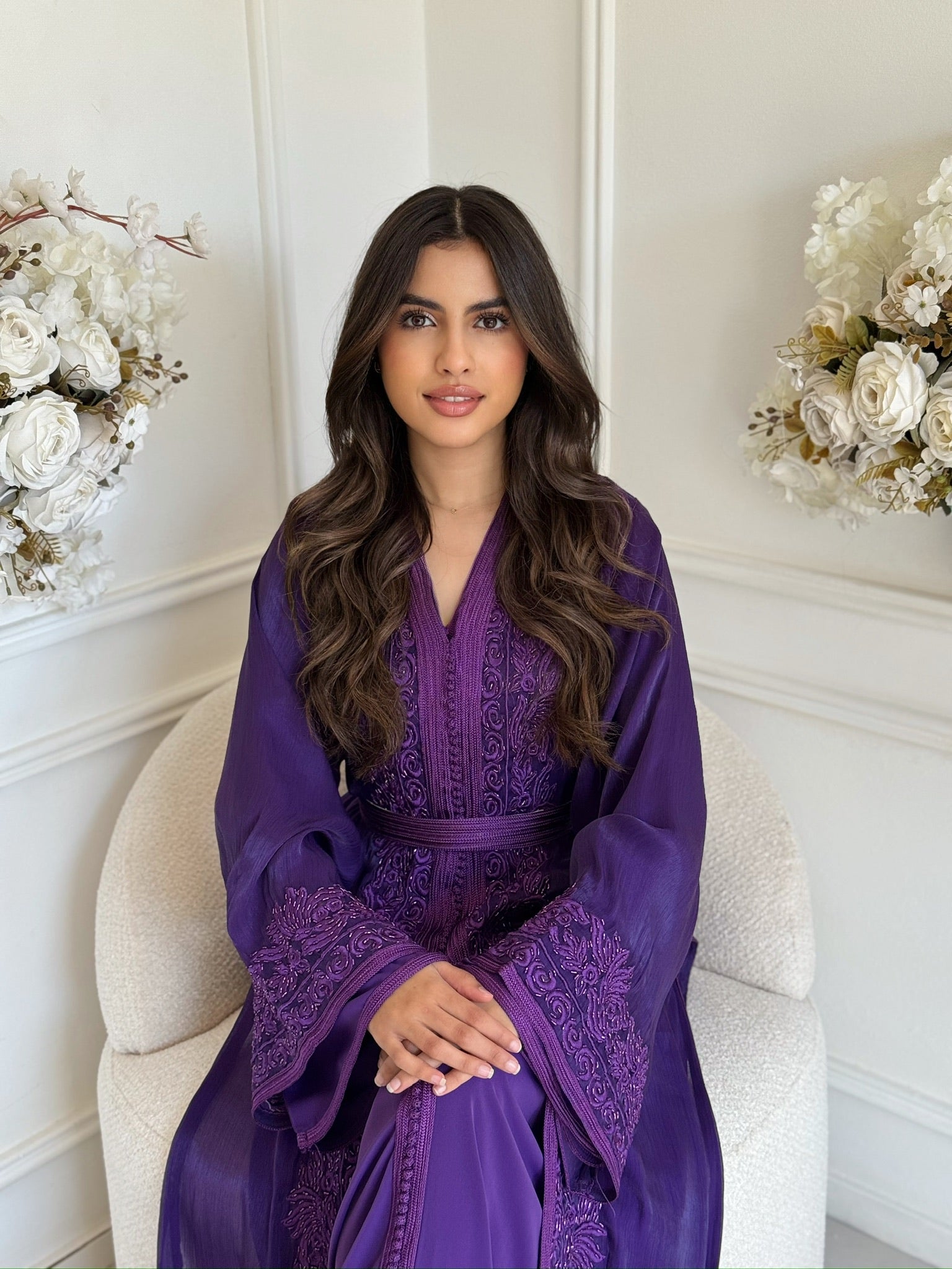 Model wearing a purple Moroccan takchita with elegant embroidery sitting indoors with white flower